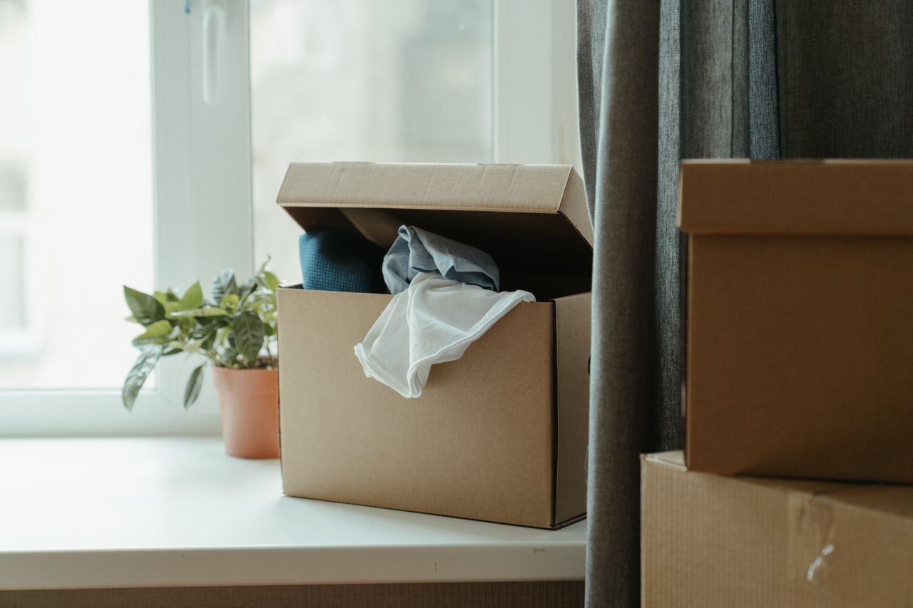 An open cardboard box filled with clothes beside a window, suggesting a move or relocation.