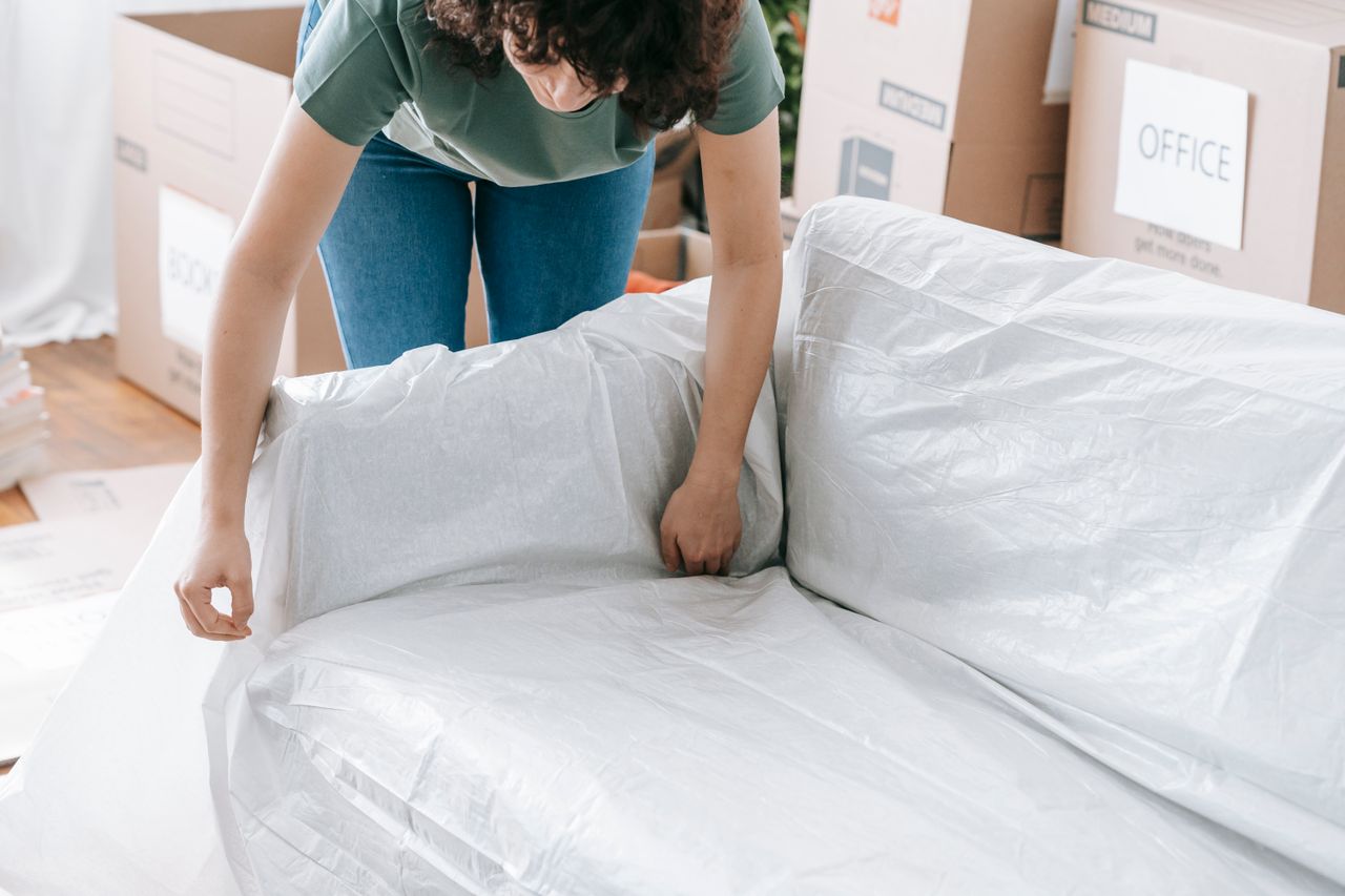 services-03 A woman gently covers a sofa with protective wrapping while packing boxes in a well-lit room.